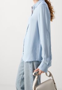 Camicia azzurra chiara a maniche lunghe con righe verticali, colletto classico e maniche con cinturino, abbinata a jeans in denim chiaro e una borsa beige.
