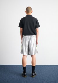 Person med rakad skalle står vänd mot en enkel vit vägg, iklädd svart tröja, silverfärgade shorts med sidoknappar, svarta strumpor och svarta skor.