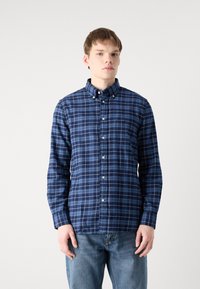 Camisa de cuadros azul marino con cuello abotonado, mangas largas y botones blancos. Presenta cuadrados en tonos azul oscuro y claro en una tela texturizada.