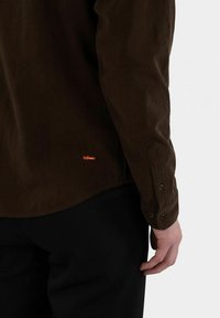 Brown corduroy shirt with long sleeves, featuring a buttoned cuff and a small orange logo near the hem. Textured fabric and tailored fit.