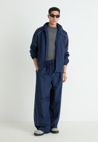 Navy tracksuit made of lightweight fabric; features a zip-up jacket with gray accents and elastic-waist tapered pants. Black shoes complete the look.