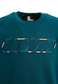 Camiseta de color verde azulado hecha de algodón, con un destacado logo bordado "MWM" en tonos plateados, cuello redondo y mangas cortas.