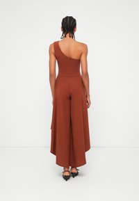 Jumpsuit u rust boji s dizajnom na jedno rame, širokim nogavicama i zatvaračem na leđima. Glatka tkanina s lepršavim siluetom. Crna obuća vidljiva.