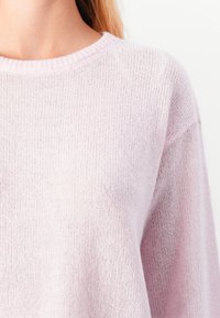 Woman wearing a soft light pink knitted sweater with a round neckline, shown from neck to mid-torso against a white background.