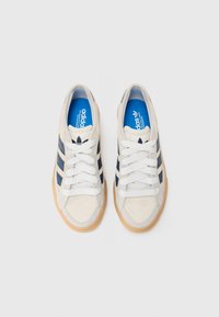 Bele tenisice iz semiša bele barve z modrimi poudarki in tremi temno modrimi črtami. Imajo bele vezalke in gumijast podplat. Vključujejo modro blagovno znamko na vložku.