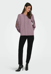 Naine kannab lillakast nööbitud pluusi pitsist varrukatega, musti kitsaid pükse ja musti kõrge kontsaga poolsaapakesi, seisab käed taskutes.