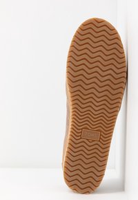 Semelle d'une chaussure TOMS marron clair avec motif de bande de roulement ondulé et logo rectangulaire au centre, présentée sur un fond blanc.