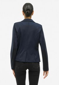 Námořnický modrý blazer se strukturovaným designem, dlouhými rukávy a jedním rozparkem na zadní straně. Hladká látka se střihaným střihem.
