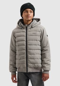 Grijze gewatteerde jas met capuchon, voorzien van een rits aan de voorkant, elastische manchetten en een logo-accent. Draagt over een donkere spijkerbroek en een zwarte beanie.