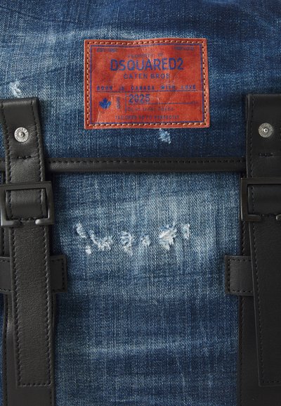 Rucsac din denim, cu un material albastru uzat, curele din piele neagră și un eticheta roșie cu text alb și un detaliu sub formă de frunză de arțar.