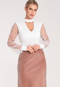 Biała bluzka z dekoltem w kształcie litery V i przezroczystymi bufiastymi rękawami ozdobionymi białymi kropkami, zestawiona z metaliczną spódnicą w kolorze różowego złota.