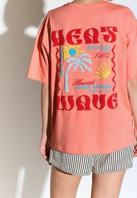 Camiseta de gran tamaño color coral con gráficos retro coloridos que presentan diseños de sol y olas, combinada con pantalones cortos a rayas en blanco y negro.