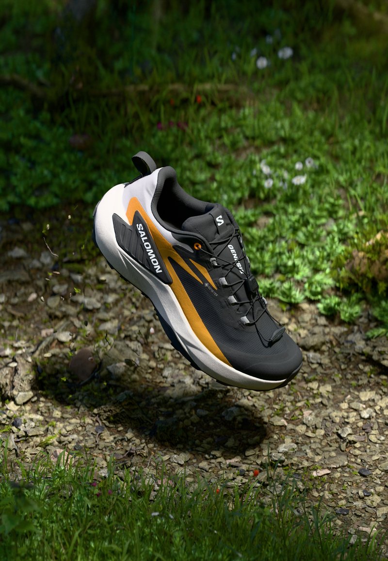 Chaussure de trail running noire et orange avec une tige texturée, un design léger et un talon de soutien, flottant au-dessus d'un sol rocailleux.