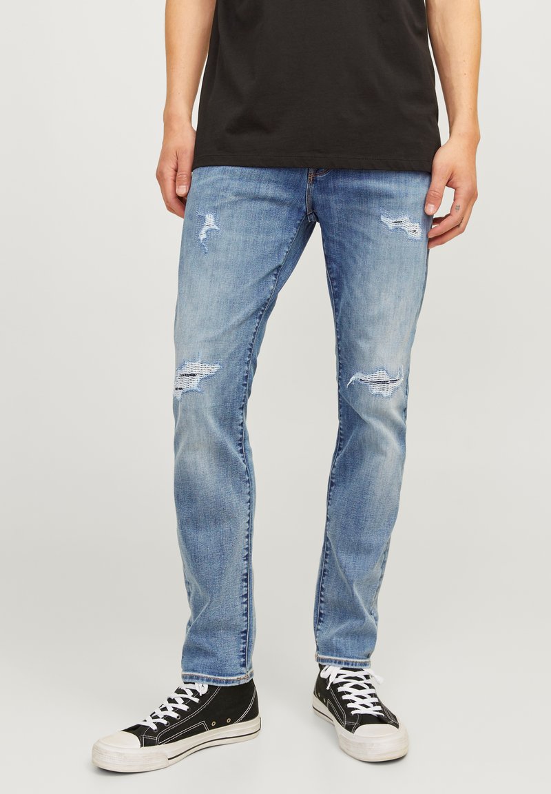 Jeans de mezclilla azul claro con parches desgastados y un corte ajustado, con un ligero desvanecimiento. Combinados con zapatillas altas negras y cordones blancos.