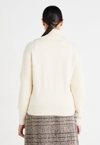 Pull crème avec un large col châle, des manches et un ourlet côtelés. La texture semble douce et tricotée, vue de dos.