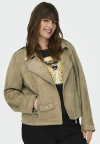 Chaqueta de motorista de ante sintético en color verde oliva con cuello acanalado, cremalleras plateadas y bolsillos laterales, combinada con una camiseta gráfica negra.