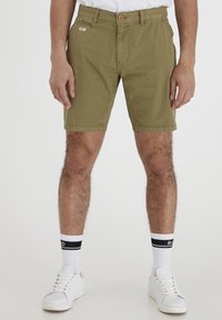 Man wearing olive green patterned shorts, white crew socks with black stripes, and white sneakers, standing against a plain background.