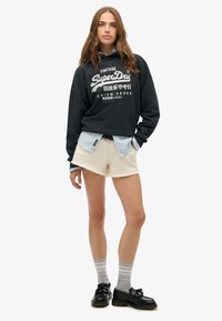 Sudadera corta negra con texto impreso en blanco, camisa de botones azul claro debajo, pantalones cortos crema, calcetines a rayas grises y zapatos negros.
