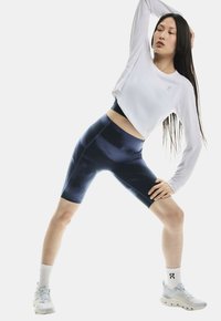 Donna in un crop top a maniche lunghe bianco e pantaloni biker blu stretchy, che si allunga di lato con un braccio alzato, indossando scarpe da ginnastica bianche e calzini.