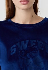 Zbliżenie na osobę w niebieskiej aksamitnej bluzie z wytłoczonym misiem i napisem "SWEET" na piersi.