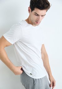 T-shirt en coton blanc avec un col rond, des manches courtes et un logo de crocodile vert en bas à gauche, associé à un pantalon de survêtement gris.