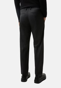 Pantalon ajusté gris foncé avec une texture lisse, devant plat et deux poches arrières. Associé à des chaussures noires élégantes.