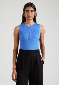 Vrouw in een mouwloos blauw geribbeld topje met knopen aan de voorkant en zwarte hooggerokte wijde broek, staande tegen een effen achtergrond.