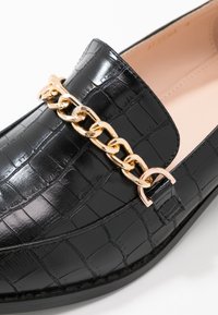 Mocassino in pelle nera con una texture in pelle di coccodrillo, caratterizzato da un accento in catena dorata e una soletta rosa liscia. Forma arrotondata con suola piatta.
