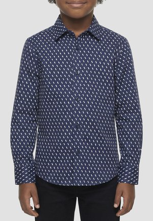 Bambino che indossa una camicia blu navy con bottoni e motivo diagonale tratteggiato bianco, pantaloni neri, in piedi con le braccia rilassate.