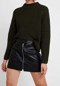 Pullover a coste di un verde scuro con collo alto, abbinato a una mini gonna in pelle nera con zip frontale e chiusura a bottone a scatto.