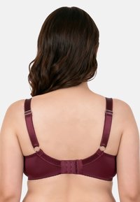 Žena s dlouhými hnědými vlasy v tmavě červené podprsence, pohled zezadu, kde jsou vidět nastavitelné ramínka a zapínání na háčky.