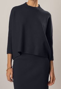 Maglione affollato di colore blu navy con collo a imbuto e orlo a coste. Caratterizzato da maniche ampie e una texture liscia, abbinato a un vestito aderente blu navy.