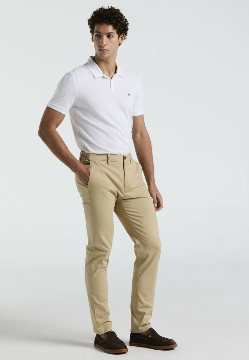Polo shirt blanc à manches courtes avec un col, pantalon chino beige et chaussures à enfiler marron foncé. Tissu lisse, coupe ajustée et design épuré.