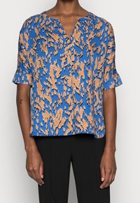 Blouse à manches courtes en tissu bleu avec un motif abstrait marron et beige. Dispose d'un col en V et d'une coupe décontractée. Texture douce.