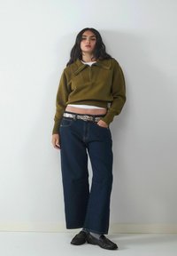 Olivgrüner, cropped Sweater mit Reißverschluss-Kragen und gefälteltem Detail. Kombiniert mit lässigen blauen Jeans und einem silberfarbenen Gürtel.