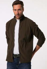 Giacca in pile marrone scuro con zip frontale, colletto alto e polsini a costine. Indossata sopra una polo bianca con tre bottoni.