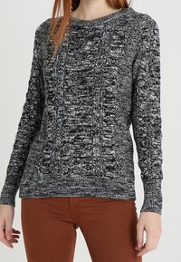 Frau, die einen schwarz-weißen melierten Strickpullover mit Zopfmustern und braunen Hosen trägt, steht vor einem einfarbigen Hintergrund.