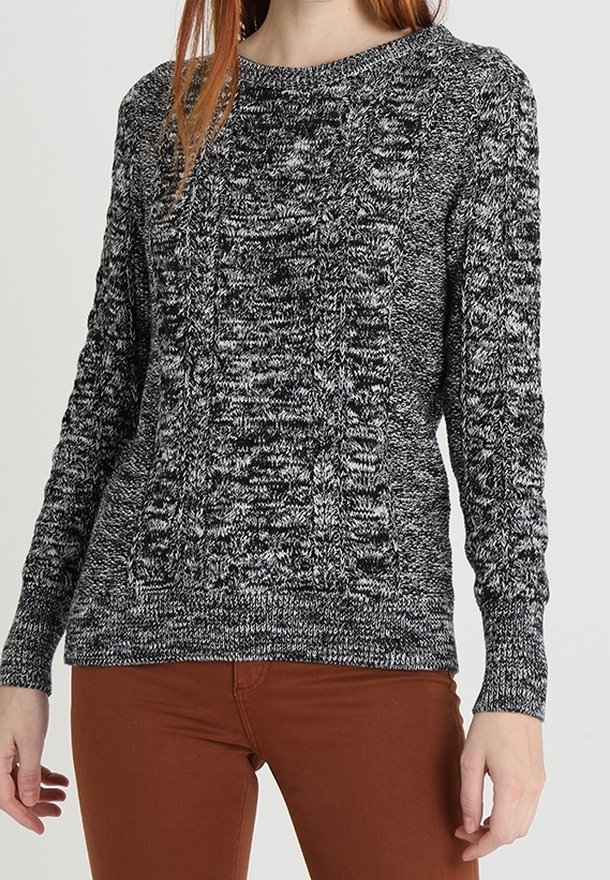 Frau, die einen schwarz-weißen melierten Strickpullover mit Zopfmustern und braunen Hosen trägt, steht vor einem einfarbigen Hintergrund.