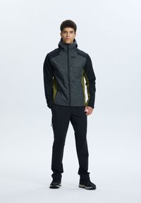 La giacca da uomo presenta un mix di maglia grigia e pannelli neri, con un design aderente e cappuccio. Abbinata a pantaloni neri e calzature scure.