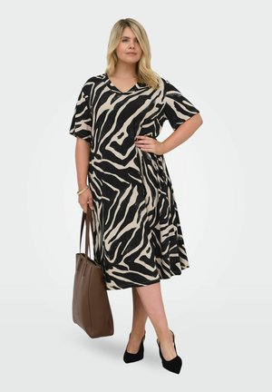 Kobieta w czarno-beżowej sukience w zebra print, czarne szpilki, trzyma dużą brązową torbę, stoi z jedną ręką na biodrze.