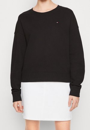 Personne portant un pull à manches longues noir avec un petit logo sur la poitrine et une jupe blanche devant un fond uni.