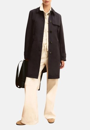 Donna con cappotto nero con bottoni, tuta color crema, scarpe beige e che tiene una borsa nera su uno sfondo semplice.