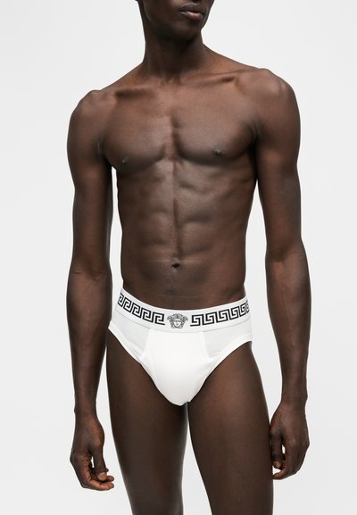 Mannequin masculin porte un slip blanc Versace avec ceinture noire à motif grec et logo Méduse, debout devant un fond blanc uni.