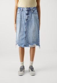 Falda midi de mezclilla en azul claro con dobladillo deshilachado, un ligero desvanecimiento y detalles desgastados. Combinada con zapatillas grises y calcetines de colores claros.