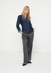 Camisa de ganga azul escura com fecho de botões e mangas dobradas, combinada com calças largas cinzentas e sapatos pretos de bico fino. Design simples e ajustado.