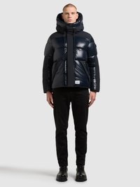 Temno modra puhasta jakna z visokim ovratnikom, zadrgo spredaj in vrvico za zategovanje. Oblečena je v sijočo teksturo in ima logotip na sprednji strani. Črne hlače in škornji.