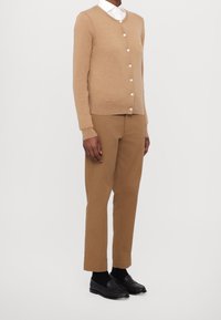 Světle hnědý vlněný cardigan s kulatým výstřihem a zapínáním na knoflíky, doplněný o hnědé kalhoty a černé loafers. Pod tím je bílá košile.