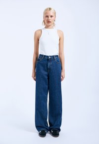 Weite blaue Denim-Jeans mit hoher Taille, akzentuiert durch fünf Taschen und einen Frontknopf. Kombiniert mit einem figurbetonten weißen ärmellosen Oberteil.