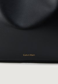 Svart lær lommebok med en jevn tekstur, subtil kurve, og gullprent "Calvin Klein" logo på bunnen av kanten.