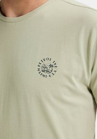 Hellgrünes T-Shirt aus weicher Baumwolle, mit rundem Halsausschnitt und einem kreisförmigen navyblauen Grafikdesign mit Text und Palmenmotiv auf der Brust.